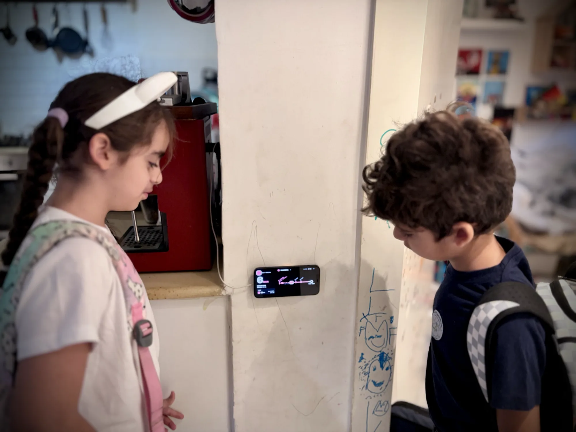 Two children with school bags in a kitchen, looking at an old iPhone mounted sideways on the wall. It shows the My Bus Stop board: the next bus in large numbers beside the line's route.
